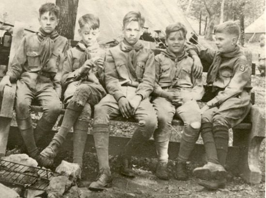An old sepia photograph of five young Scouts in uniform and neckers, sitting together on a bench at camp